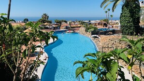 A pool with a curved design, surrounded by palm trees and a view of the sea.