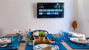 A dining table set with blue placemats, plates, cutlery, and glasses, with a TV mounted on the wall displaying various streaming services. - Vacation Home GIRO DI BITTA (Siculiana Marina)
