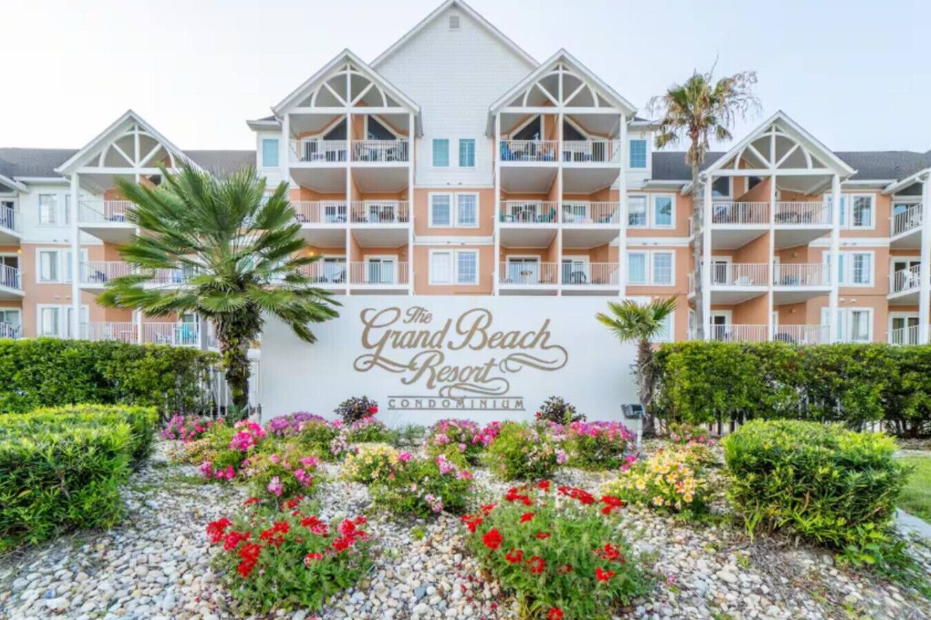 Ein mehrgeschossiges Gebäude mit Balkonen, einem Schild mit der Aufschrift 'The Grand Beach Resort Condominium' und gepflegten Gärten mit verschiedenen Pflanzen und Blumen.