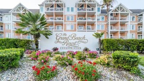 Un immeuble à plusieurs étages doté de balcons, une enseigne indiquant « The Grand Beach Resort Condominium », et des jardins aménagés avec différentes plantes et fleurs.