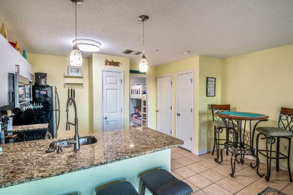 A kitchen with granite countertops, a refrigerator, a sink, and a dining area with chairs and a table. - Whale of a Time! Grand Beach (Gulf Shores)
