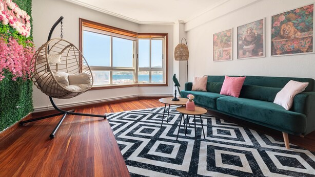 Un salon moderne avec un canapé vert, un fauteuil suspendu, un tapis à motifs géométriques et une grande fenêtre offrant une vue dégagée.