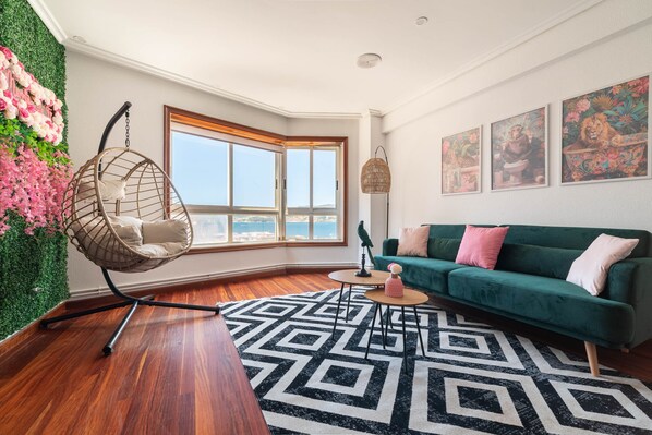 A modern living room with a green sofa, a hanging chair, a geometric rug, and a large window with a view.