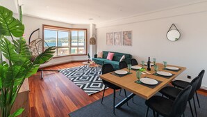 Un salon moderne avec une table à manger en bois, des chaises noires, un canapé vert et une grande fenêtre donnant sur la mer.