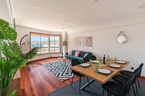 A modern living room with a wooden dining table, black chairs, a green sofa, and a large window offering a view of the sea.