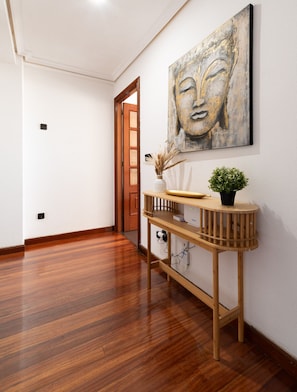A hallway with wooden flooring, a wooden console table with a vase and plant, and a large abstract painting on the wall.