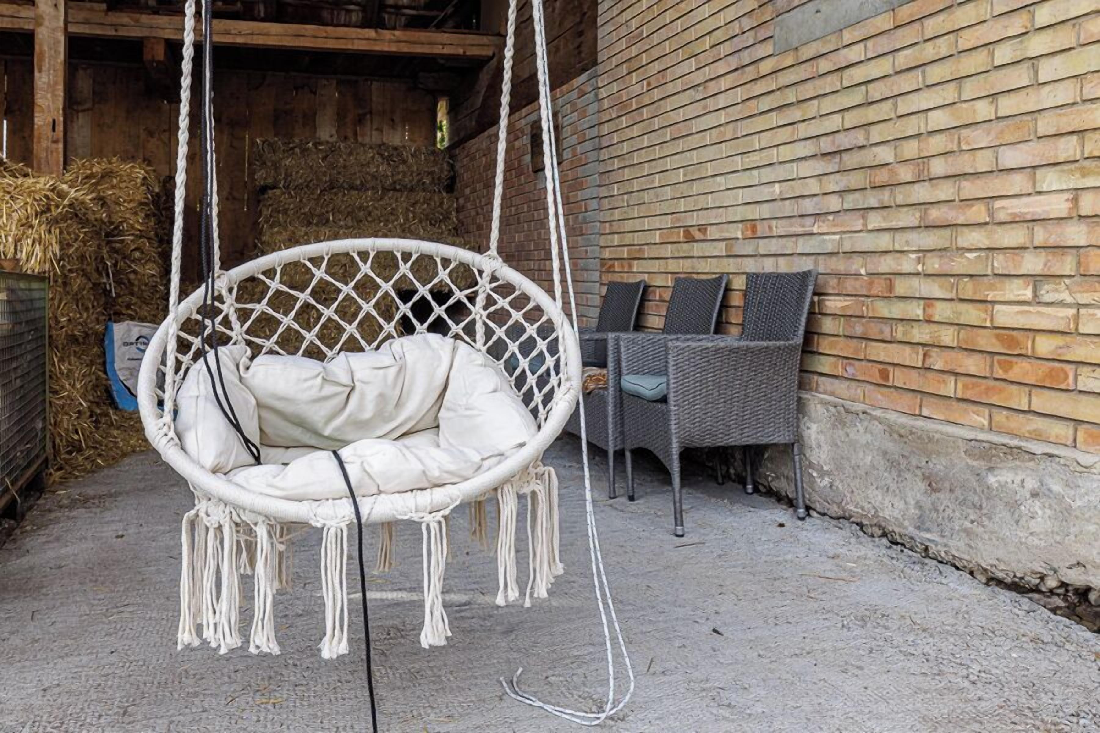 A white hanging chair with a cushion, a wicker chair with cushions, and a brick wall in the background.