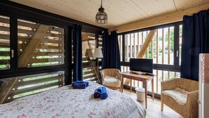 Une pièce de style cabane en bois, avec un lit, deux fauteuils en osier, une petite table avec un téléviseur et de grandes fenêtres.