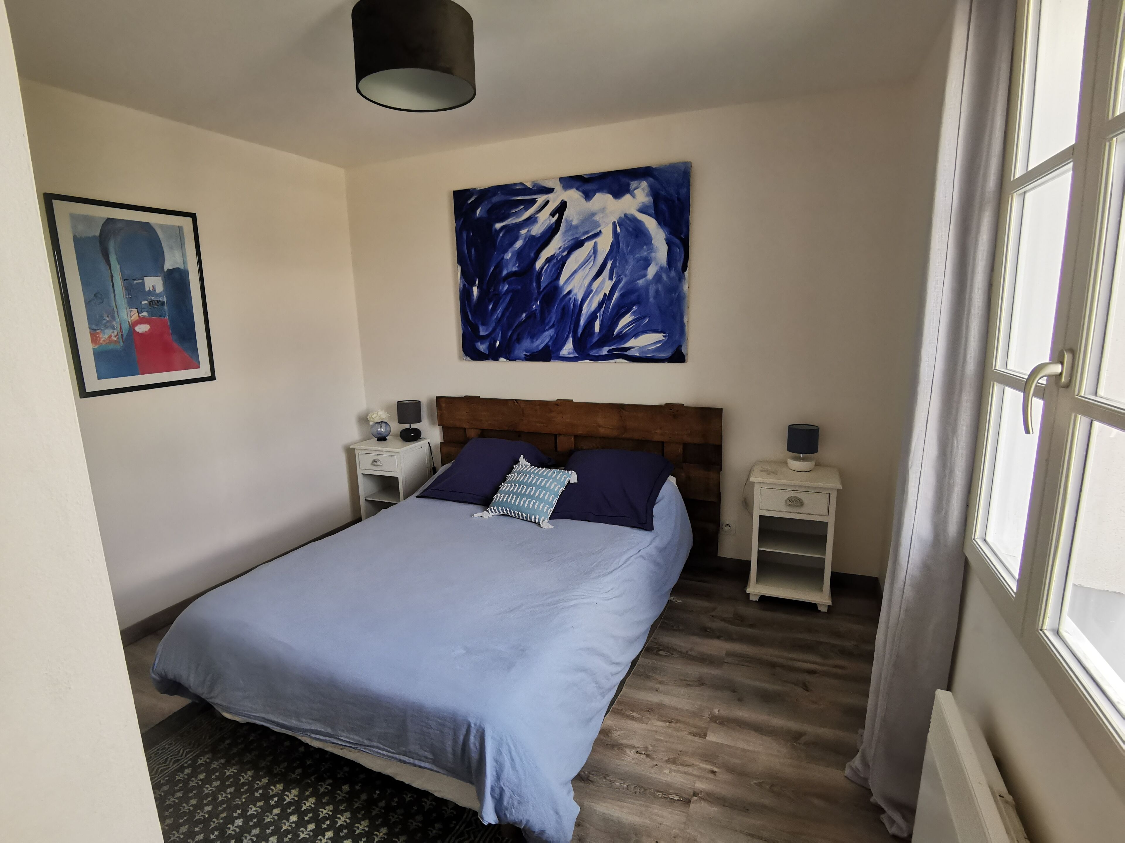 A bedroom with a bed, two bedside tables, a nightstand, a painting, and a window.