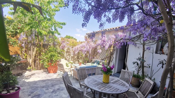 A patio with a round wooden table and chairs, surrounded by potted plants and purple flowering trees. - Village house with garden. Pets allowed (La Flotte)
