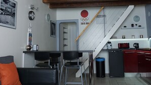 A modern living space with a wooden ceiling, a black sofa, a wooden table, a kitchen area with a sink and cabinets, and a staircase.