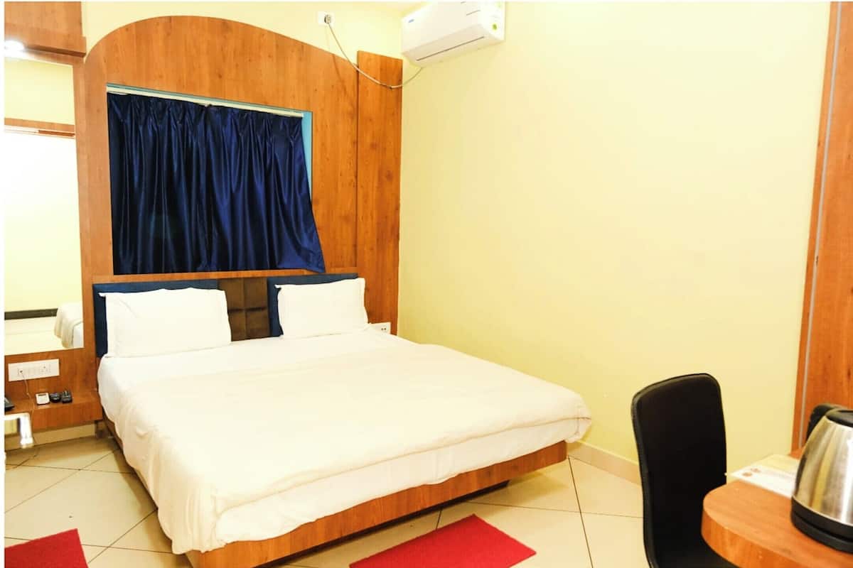 A hotel room with a wooden bed frame, a large window with a blue curtain, a small table with a kettle, and a chair.