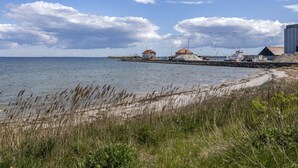 A coastal scene with a sandy beach, grassy dunes, and a body of water. There are buildings with red roofs and a large industrial structure on the right. - "Ilze" - 20m from the sea by Interhome (Bandholm)