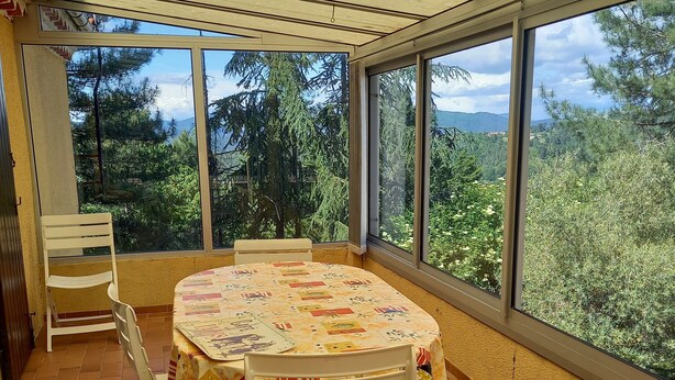 A covered patio with a round table and chairs, large windows, and a view of trees and hills. - Renovated apartment in quiet country house with view of the Cevennes mountains (Portes)