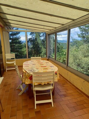 Eine überdachte Terrasse mit rundem Tisch und Stühlen, großen Fenstern und Blick auf Bäume und Hügel.