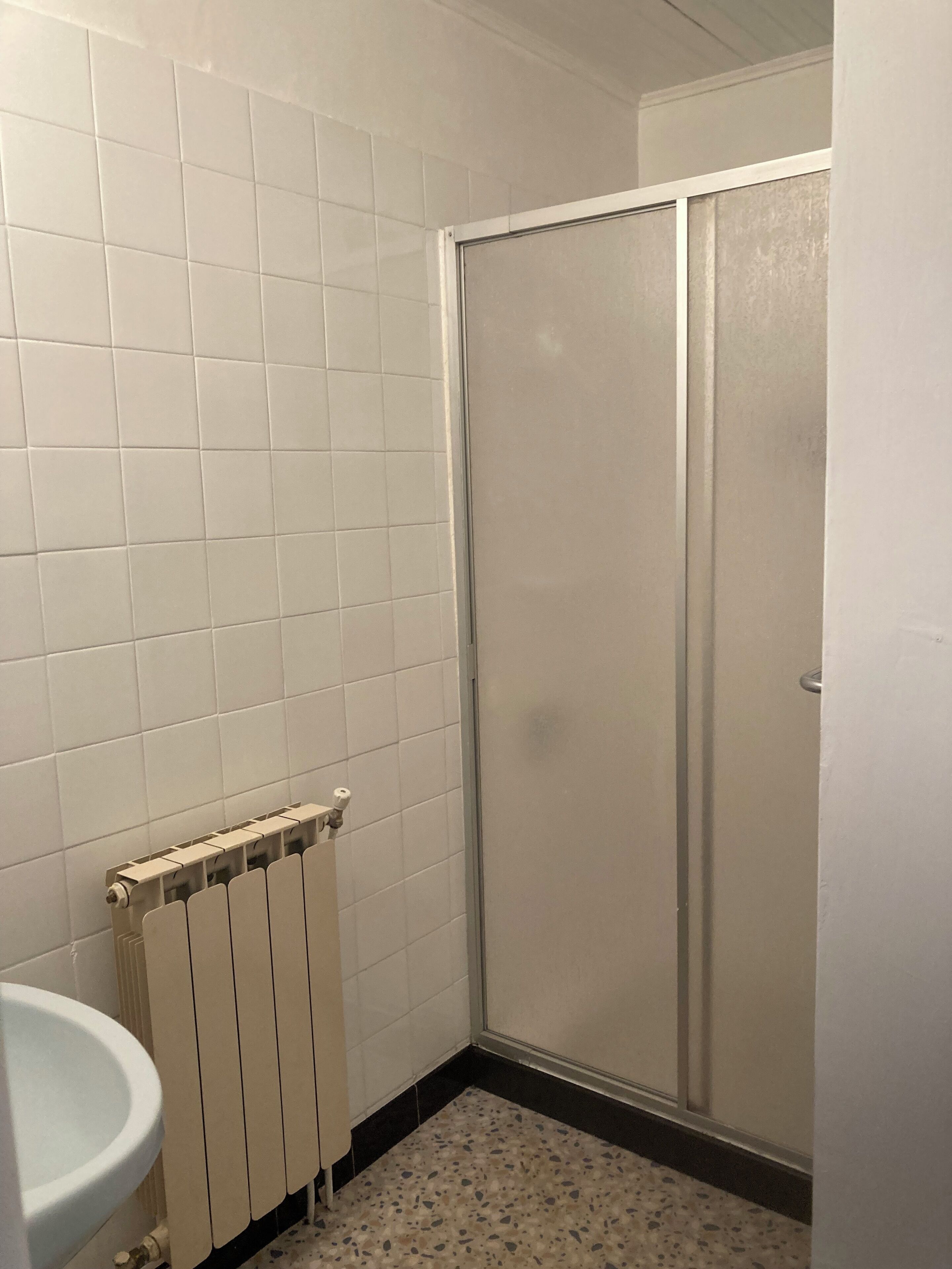 A bathroom with white tiles, a white sink, a radiator, and a shower with a glass door.