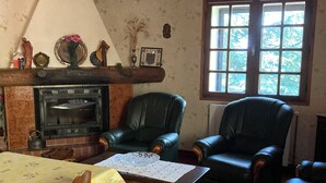 A cozy living room with a fireplace, leather chairs, a dining table with a floral tablecloth, and a large window with a view of trees. - Renovated apartment in quiet country house with view of the Cevennes mountains (Portes)