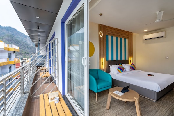 A balcony with a glass door, a bed with a striped headboard, a teal armchair, and a small wooden table. - goSTOPS PLUS Rishikesh, Tapovan (Rishikesh)