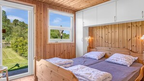 A bedroom with wooden walls and ceiling, a large window with a view of a garden, a bed with two pillows, and a bedside table.