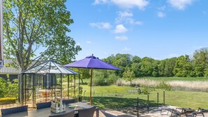 A wooden deck with outdoor furniture, a glass gazebo, and a view of a grassy area with trees.
