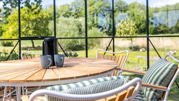 A round wooden table with two mugs, surrounded by white wicker chairs with striped cushions, under a glass greenhouse structure with a view of greenery outside.