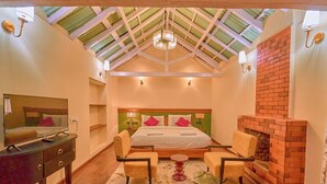 A bedroom with a large bed, two chairs, a small table, a TV on a dresser, and a brick fireplace. - Mowbray House Resort Ooty by VOYE HOMES (Ooty)