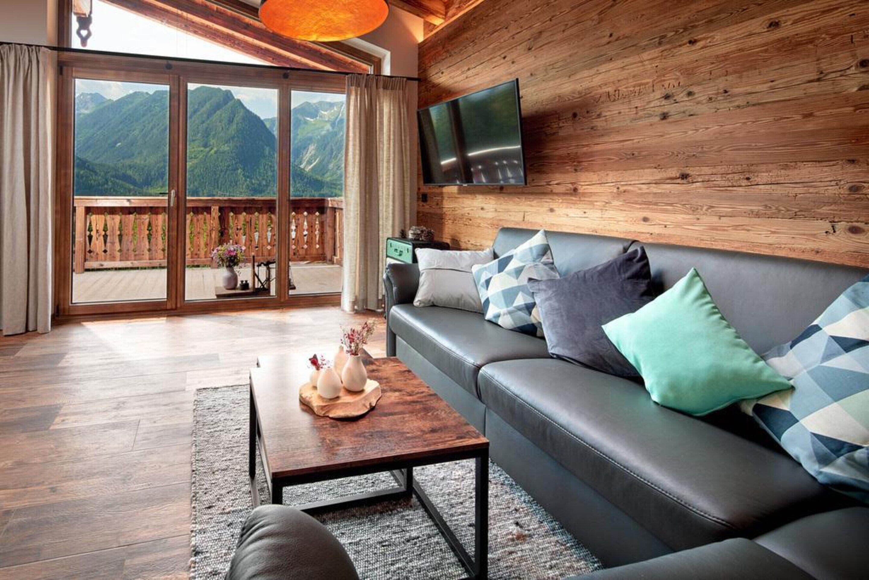 A modern living room with a wooden wall, a leather sofa, a coffee table with decorative items, and a large sliding glass door leading to a balcony with a mountain view.