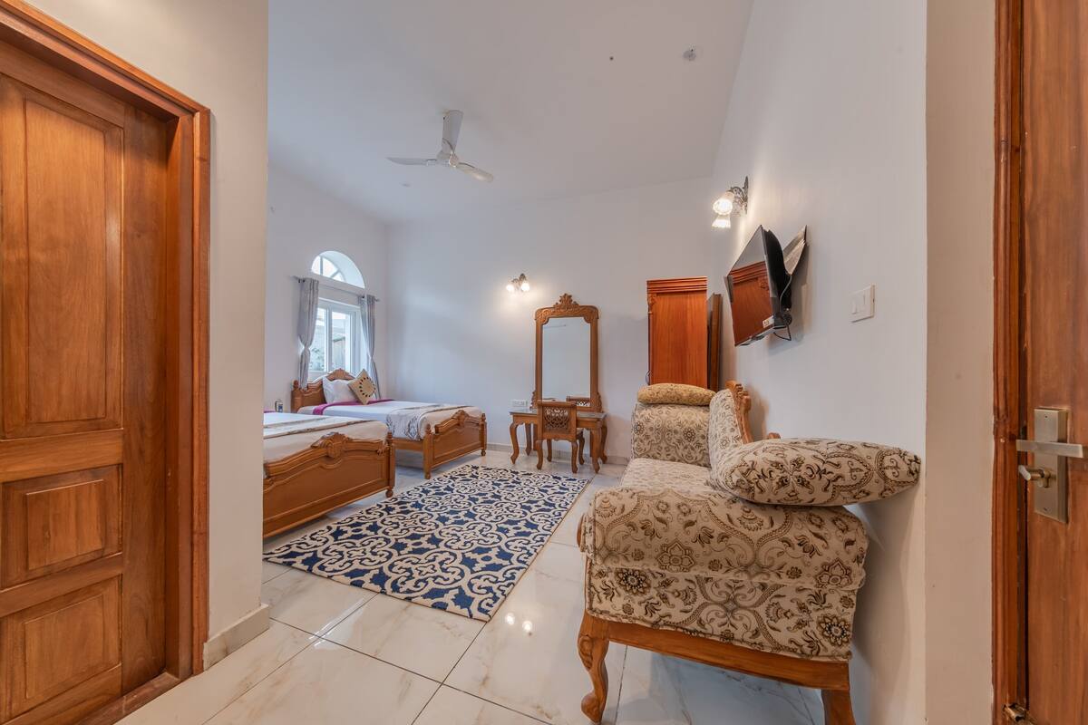 Un dormitorio con puerta de madera, cama con alfombra azul estampada, una silla de madera, un espejo y un ventilador de techo.