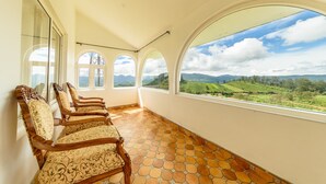 Ein Zimmer mit Fliesenboden, Holzstühlen und großen Bogenfenstern mit Blick auf grüne Hügel.