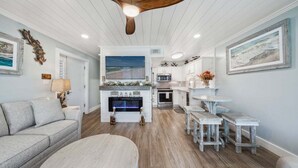 Un salon chaleureux avec une cheminée, un coin repas avec des bancs en bois et une cuisine avec des meubles blancs.