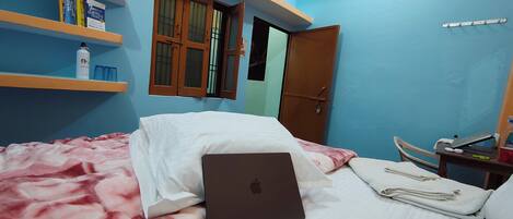 A bedroom with a bed, a laptop, a nightstand, and a shelf with books and a bottle.