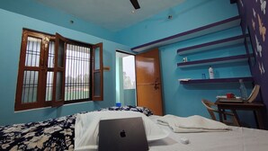 A bedroom with a bed, a laptop, a ceiling fan, a window with bars, a door, and a shelf with toiletries. - Sneh Homestay (Faizabad)