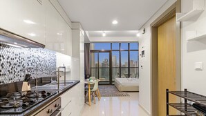 A modern kitchen with a gas stove, a dining area with a table and chairs, a large window with a city view, and a bedroom with a bed and a nightstand. - FIRST CLASS | Studio | Chic City Escape (Dubai)