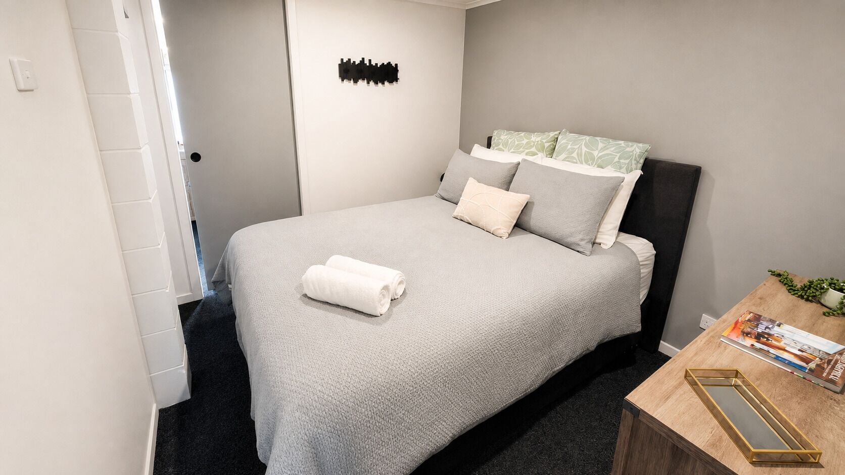A neatly made bed with a grey bedspread and pillows, a small bedside table with a book and a tray, and a wall-mounted light switch.