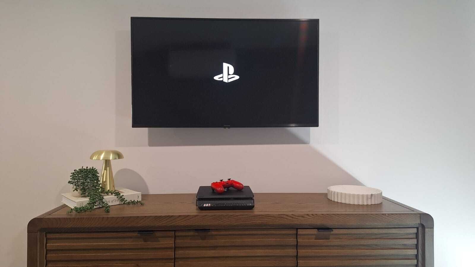 Een flatscreen televisie bevestigd aan een muur boven een houten kastje met een decoratieve lamp, een kleine plant en een rood object.