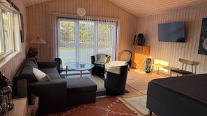 Ein gemütliches Zimmer mit einer Couch, einem kleinen Tisch, einem Sessel, einem Fernseher und einem Fenster mit Jalousien.