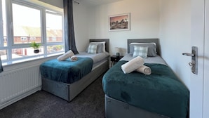 Une chambre avec deux lits, une fenêtre avec des rideaux, un radiateur, une table de chevet avec une lampe et un tableau au mur.