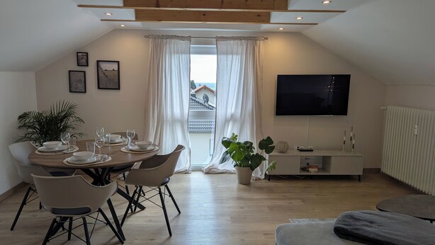 A dining area with a round table set for four, surrounded by modern chairs. A large potted plant is placed near the window, which has white curtains. A flat-screen TV is mounted on the wall above a white cabinet. - Schweigen-Rechtenbach - modern vacation apartment "Martina" ideal for four people (Schweigen-Rechtenbach)