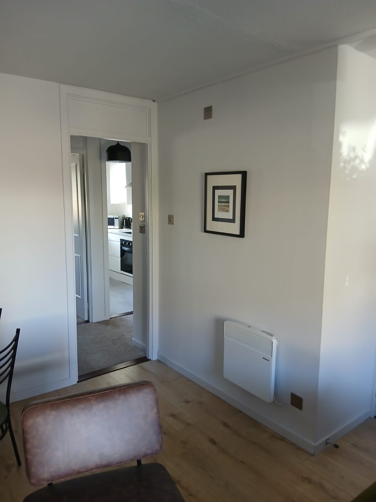 Een kamer met houten vloer, een radiator, een inlijsting foto aan de muur en een deur naar een keuken.