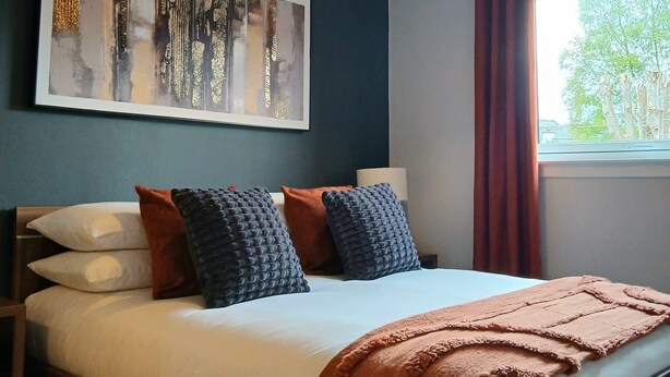 A bedroom with a bed, pillows, a blanket, a ceiling light, a window with curtains, and a wall-mounted artwork. - Newly refurbished ground floor flat within easy walking distance of town centre. (Inverness)