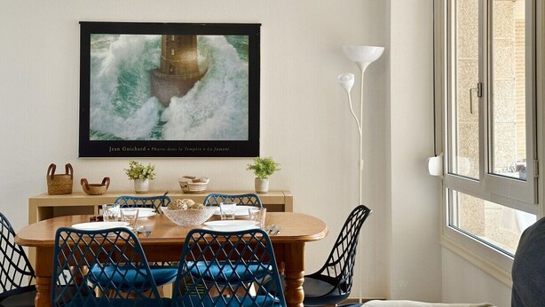 A dining area with a wooden table, blue chairs, a woven pendant light, a framed artwork, and a window with blinds. - Benvenuti 30026 Apartment in Somo (Somo)