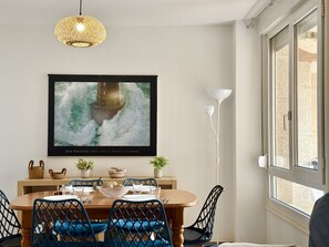 Een eethoek met een houten tafel, blauwe stoelen, een geweven hanglamp, een lijstwerk met kunst en een raam met jaloezieën.