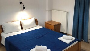 Cama bem arrumada com colcha azul, almofadas brancas e duas toalhas dobradas. Há um abajur, uma mesa de cabeceira de madeira e um radiador.