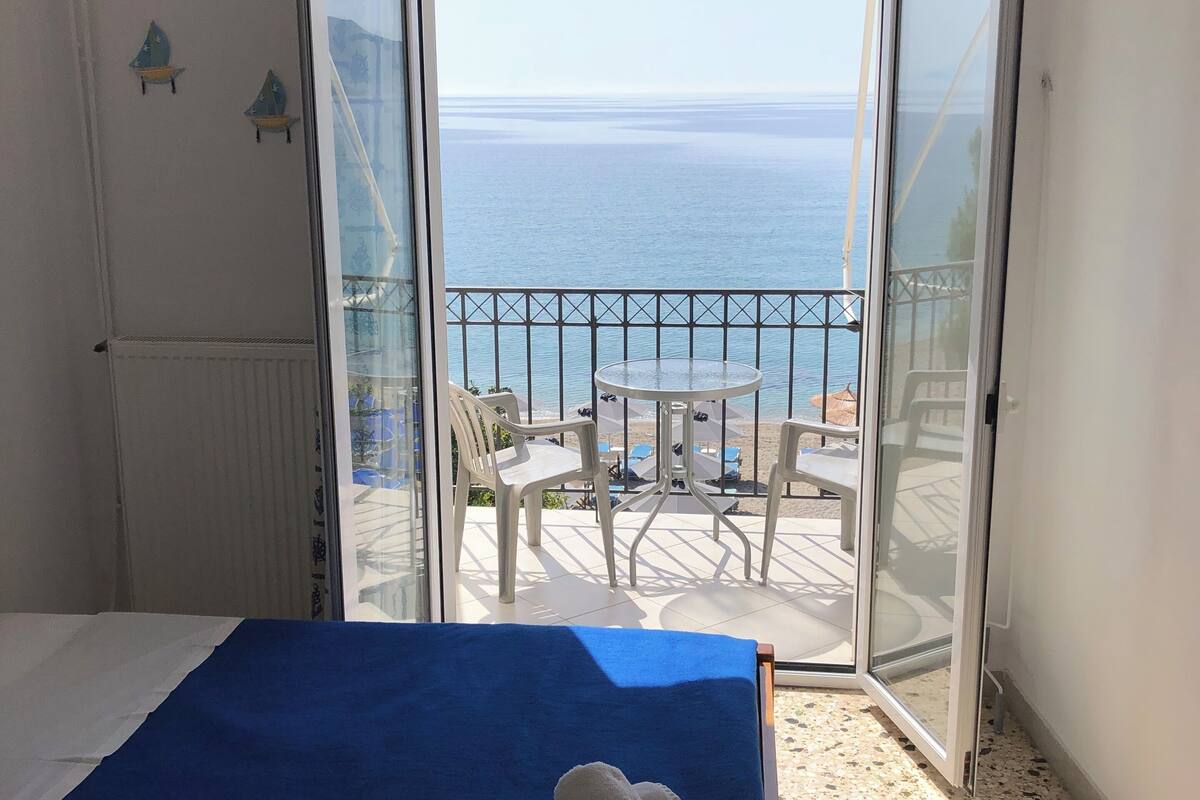 A room with a bed, a balcony with a view of the sea, and a table with chairs.