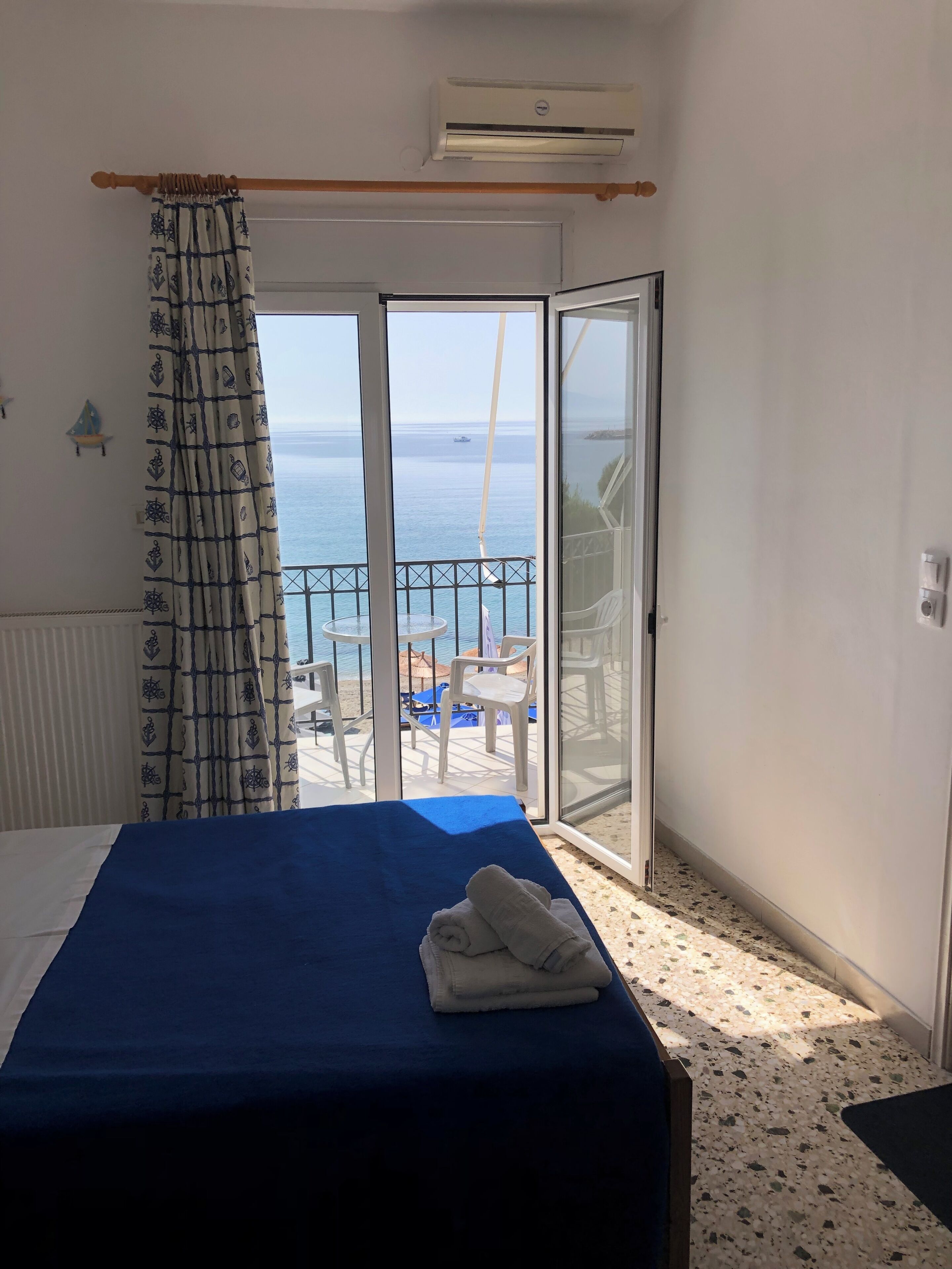 A hotel room with a bed, a curtain, an air conditioning unit, a view of the sea, and a balcony with chairs.