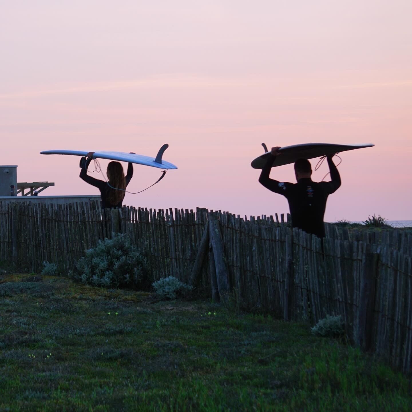 Two individuals carrying surfboards over a wooden fence.