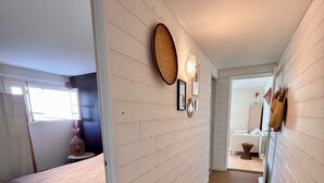 Een smalle gang met een bed aan de linkerkant, een wandlamp en een deur die naar een andere kamer leidt.
