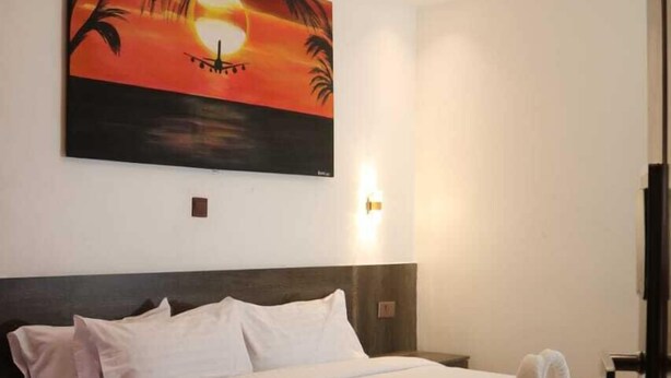 A hotel room with a bed, a painting of a sunset, a wall-mounted light, and a ceiling light fixture. - old station lounge (Naivasha)