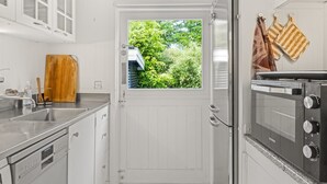 A compact kitchen with white cabinetry, a built-in oven, a window with a view of greenery, and a wooden cutting board.