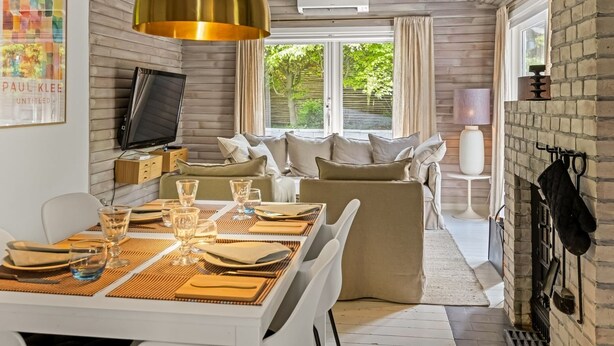 A modern dining area with a wooden table set for two, a television on a wooden cabinet, and a sofa in the background.